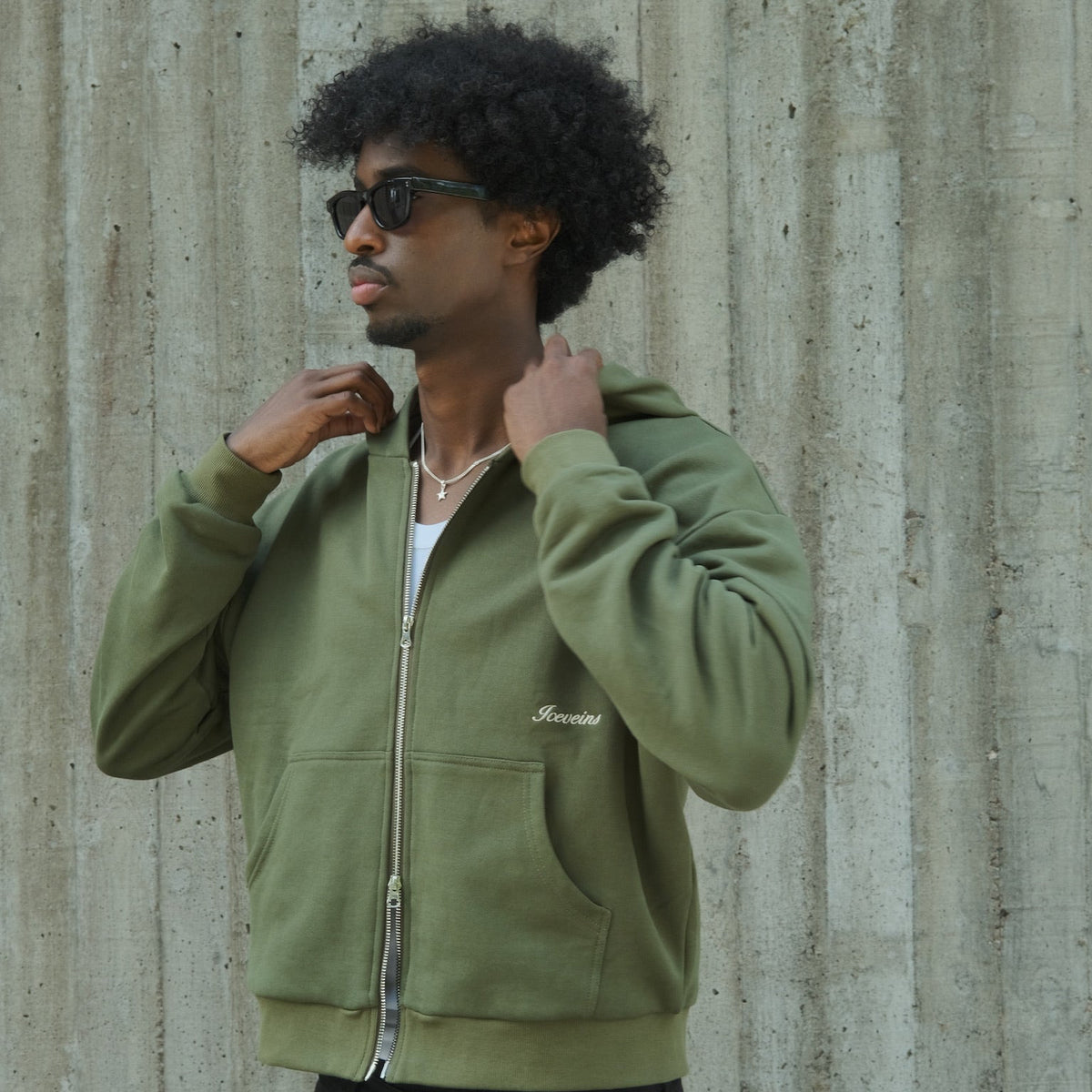 A man with curly black hair and sunglasses wearing an olive green zip-up hoodie with a small white embroidered logo, posed against a textured concrete wall.